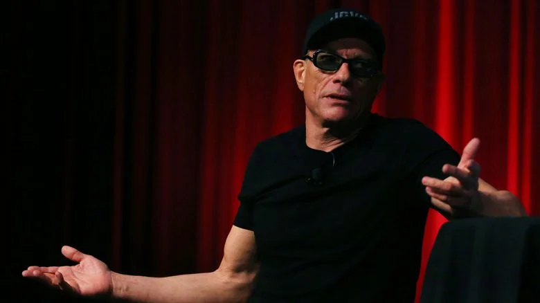 Jean-Claude Van Damme portant une casquette et des lunettes de soleil sur un fond rouge