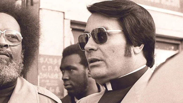 Jim Jones à un rassemblement en tenue de clergé avec des lunettes de soleil
