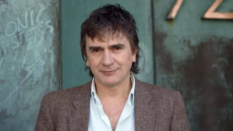 Dudley Moore en costume gris souriant devant une porte verte