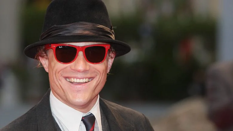Heath Ledger avec un chapeau trilby, lunettes de soleil rouges, costume, souriant