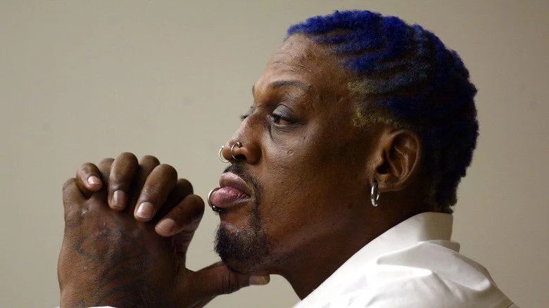Dennis Rodman avec un t-shirt blanc et des cheveux violets, les mains jointes