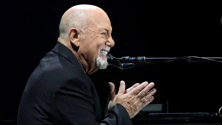 Billy Joel en costume, souriant, assis au piano