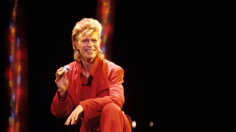 David Bowie en costume rouge, souriant et tenant un microphone