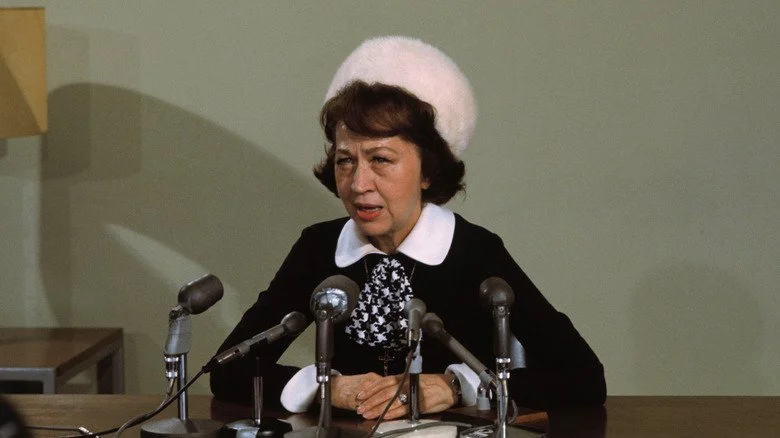 Jeane Dixon speaking at microphones, wearing a fuzzy hat