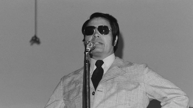 Jim Jones portant des lunettes de soleil, parlant à un microphone