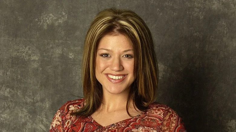 Kelly Clarkson souriante avec un fond en pierre et des reflets blonds