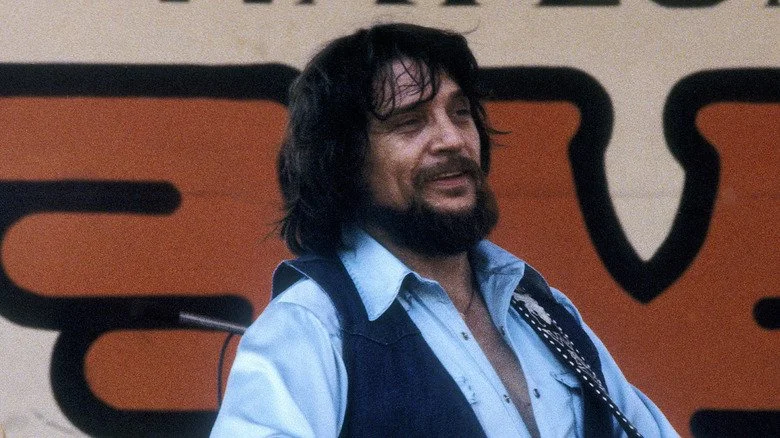 Waylon Jennings souriant en jouant de la guitare sur scène