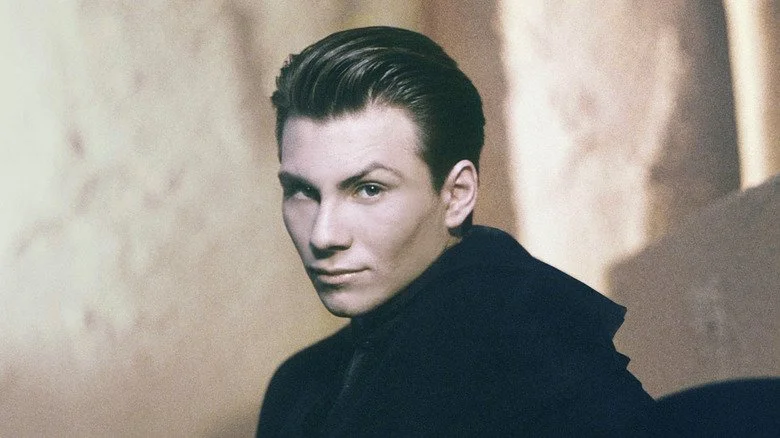 Christian Slater avec un regard espiègle et cheveux en arrière, portant une veste noire