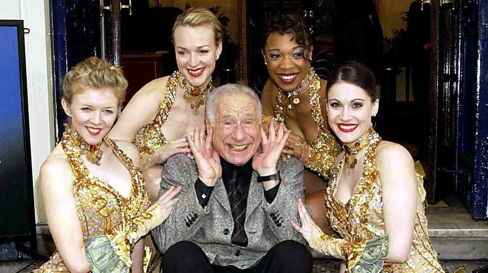 Mel Brooks souriant entouré de danseuses