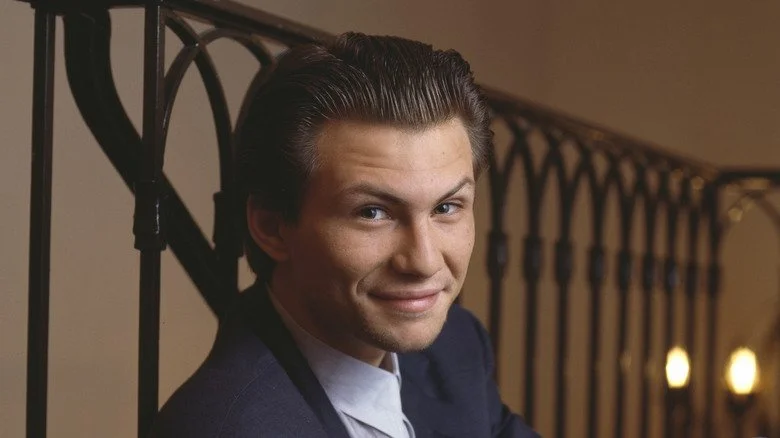 Christian Slater souriant en costume, appuyé sur une rambarde en intérieur