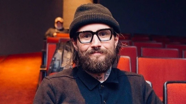 Spencer Rollins assis et souriant avec des lunettes, une barbe et un bonnet