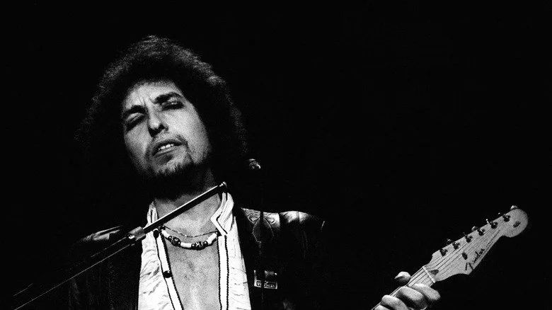 Bob Dylan on stage with leather jacket beaded necklace and guitar
