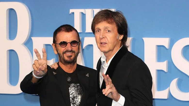 Ringo Starr faisant le signe de la paix aux côtés de Paul McCartney devant le logo des Beatles