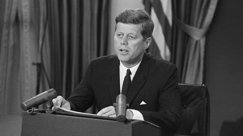 JFK assis derrière un bureau avec des microphones