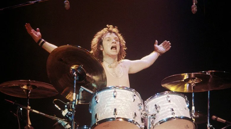 Joey Kramer shirtless and holding arms open wide at his drum kit