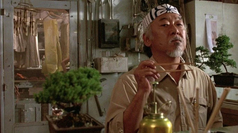 Pat Morita dans le rôle de Mr. Miyagi observant un bonsaï dans The Karate Kid (1984)