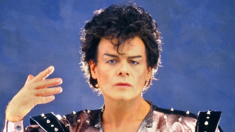 Gary Glitter en tenue glam des années 70, levant la main, fond bleu