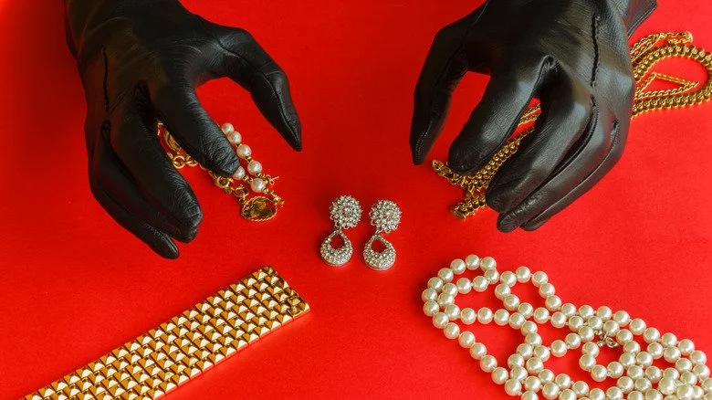 Deux mains gantées s’approchent des bijoux sur fond rouge.