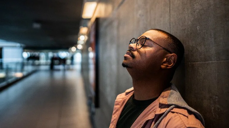 Homme portant des lunettes ferme les yeux et s'appuie contre un mur.