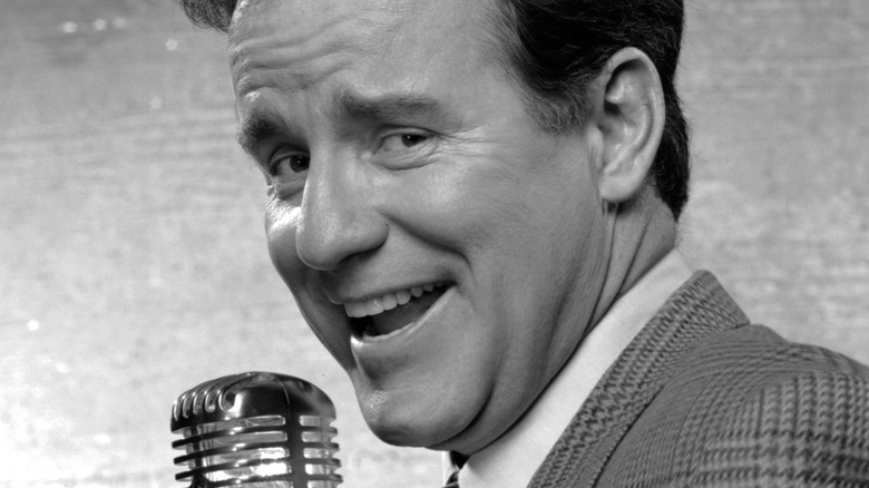 Phil Hartman en costume souriant avec un micro