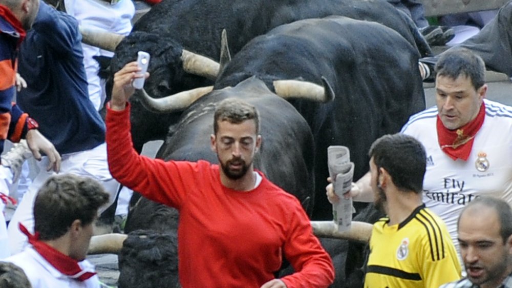 dangerous social media photo, man in a red sweater holding a mobile phone above his head while running through a crowd of people away from a charging bull