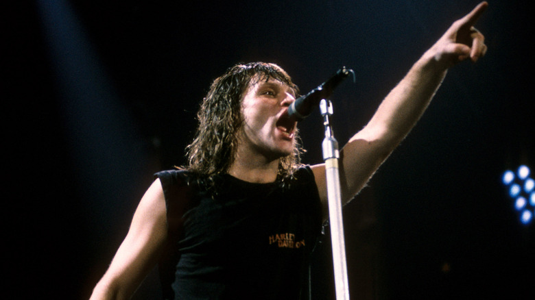 Jon Bon Jovi en chemise sombre pointant le doigt vers le haut sur scène