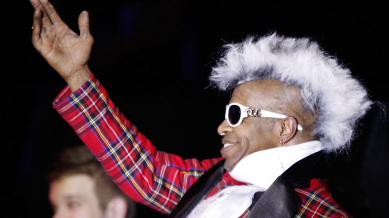 Sly Stone portant des lunettes de soleil et un costume écossais rouge, souriant avec la main levée