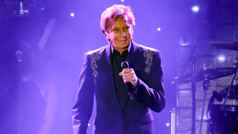Barry Manilow souriant en costume violet sur scène en 2025